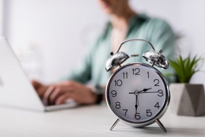 Eine Frau arbeitet an einem Laptop. Vor ihr steht ein Wecker, der auf 14:30 Uhr zeigt. Symbolbild für den steigenden Arbeitsdruck.