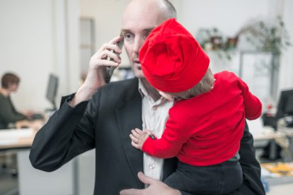 Ein Mann im Anzug telefoniert mit dem Handy arbeitet und hat dabei sein Kind auf dem Arm. Väter in Elternkarenz sind eine Minderheit.