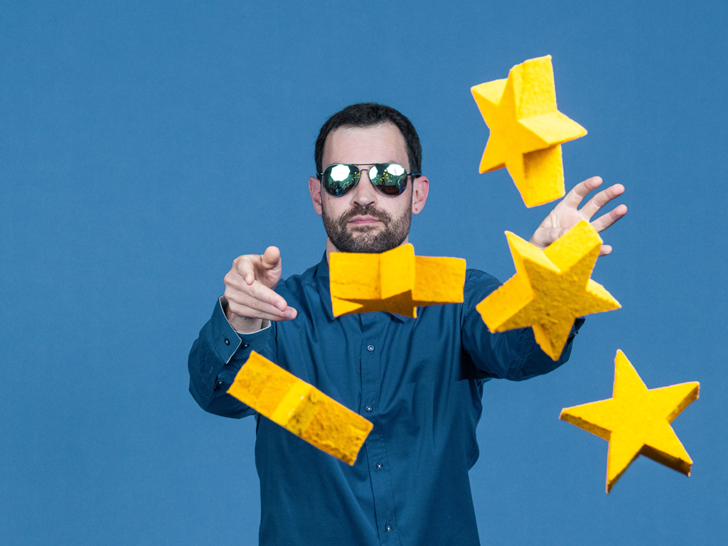 Ein Mann mit Sonnenbrille und blauem Hemd wirft gelbe Sterne in die Kamera. Symbolbild für die EU.