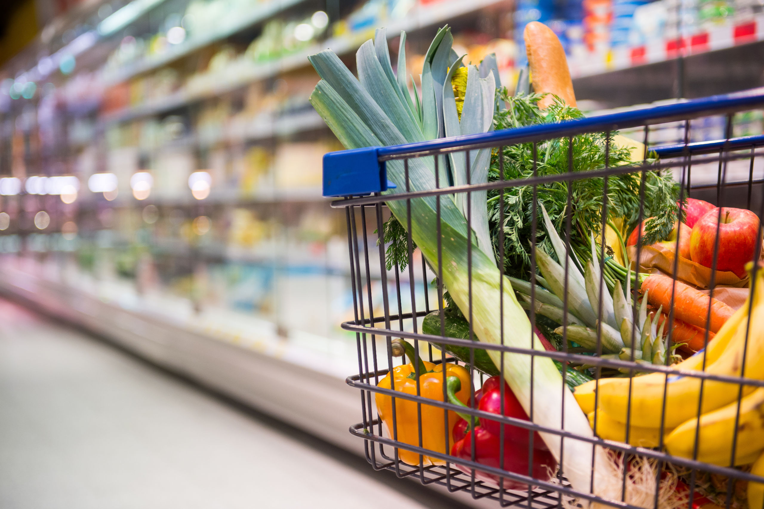 ein einkaufswagen voller Obst und Gemüste vor einem Kühlregal im Supermarkt. Die Inflation macht ihn immer teurer.