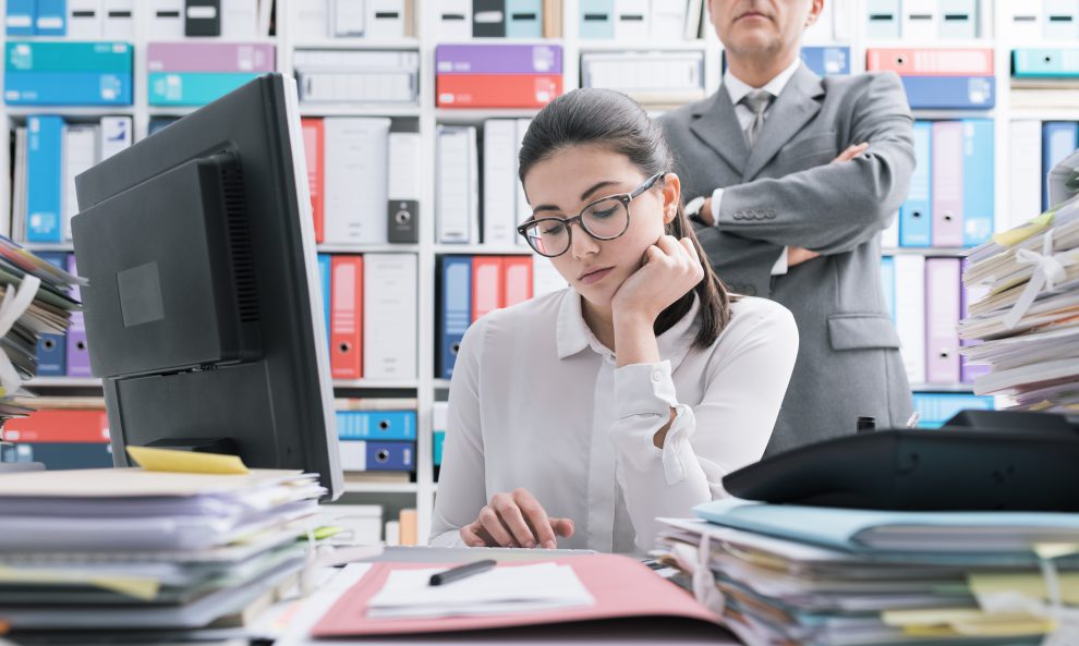 Eine junge Frau sitzt an ihrem Schreibtisch und arbeitet und wird von ihrem Chef beobachtet. Der Arbeitsklima Index zeigt, dass der Arbeitsdruck bei Lehrlingen und Auszubildenden steigt.