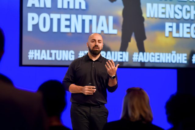 Ali Mahlodji steht als Redner auf einr Bühne.