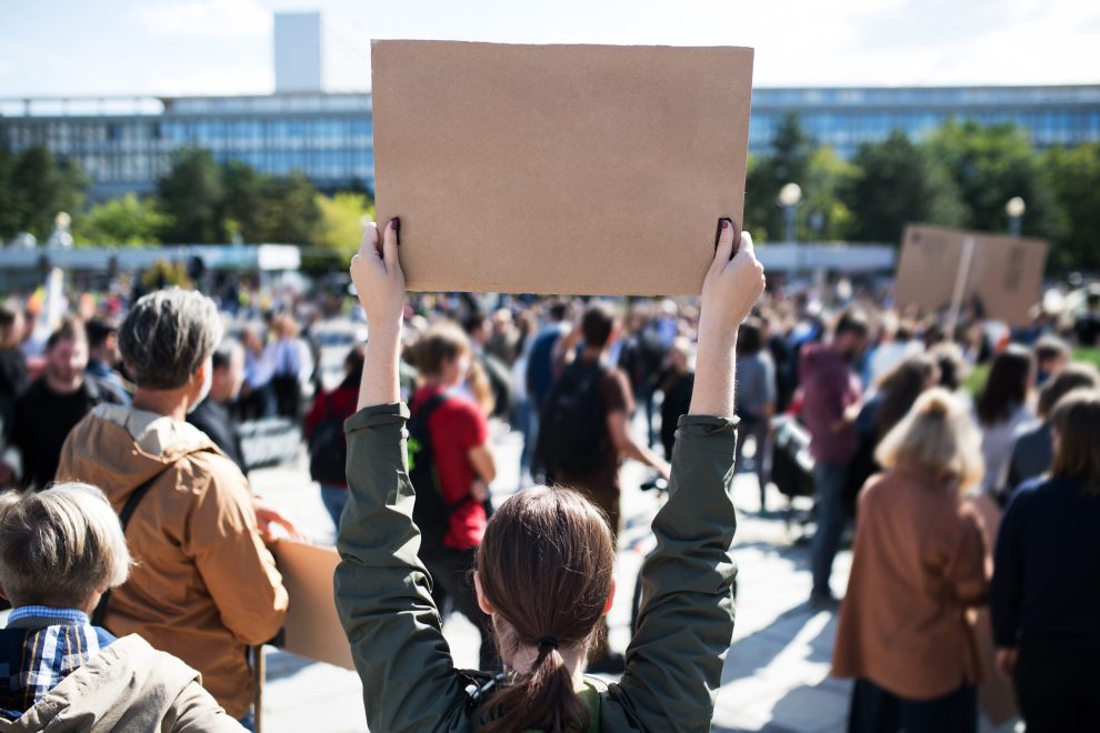 Menschen bei einer Demonstration. Eine Frau hält ein Plakat hoch. Symbolbild für den Kommentar von Kid Pex.