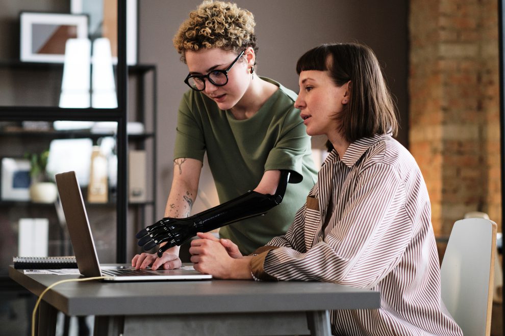 Junge Frau mit Prothesenarm, die auf einen Laptop zeigt und während der Arbeit im Büro mit ihrer Kollegin spricht. Symbolbild für Frauen mit Behinderungen und ihre Job-Suche.